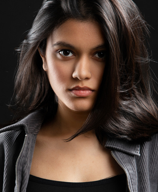 A young woman with long dark hair and a serious expression looks directly at the camera. Captured by an Ann Arbor Actor Headshots Photographer, she wears a dark zip-up jacket over a black top, set against a black background.