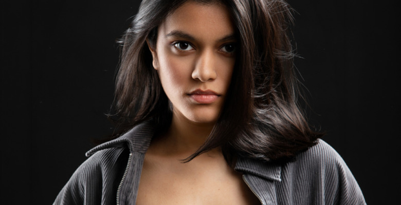 A young woman with long dark hair and a serious expression looks directly at the camera. Captured by an Ann Arbor Actor Headshots Photographer, she wears a dark zip-up jacket over a black top, set against a black background.