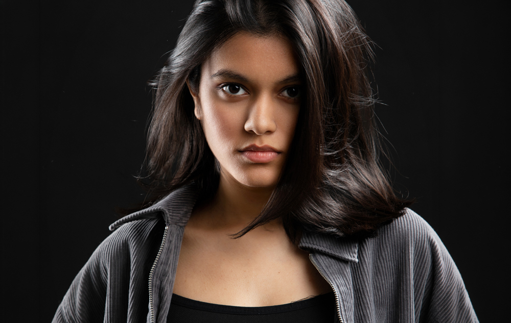 A young woman with long dark hair and a serious expression looks directly at the camera. Captured by an Ann Arbor Actor Headshots Photographer, she wears a dark zip-up jacket over a black top, set against a black background.