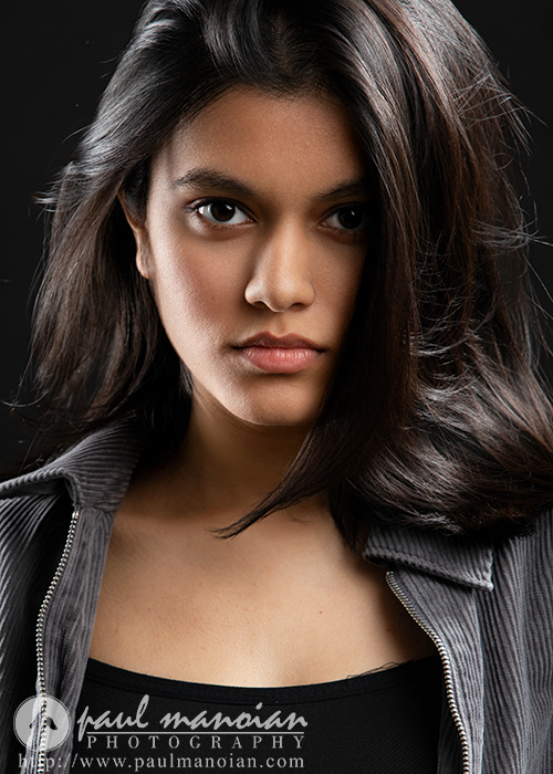 A young woman with long dark hair gazes confidently at the camera, wearing a black top and a gray-striped jacket against a dark background. Captured by Ann Arbor Actor Headshots Photographer, the image features the logo "paul manoian photography.