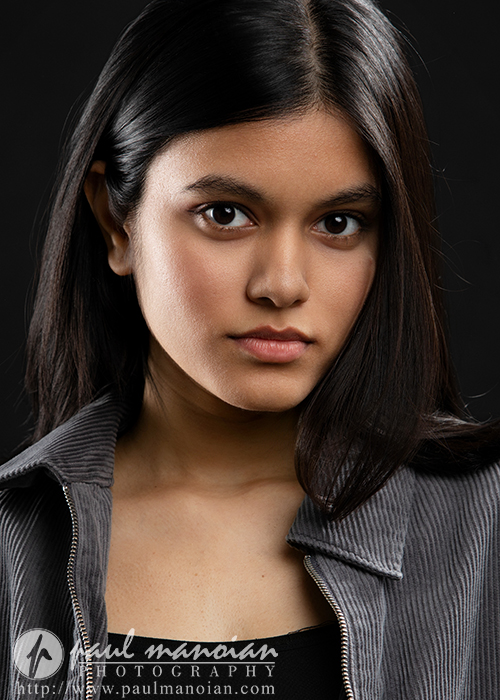 A young woman with long dark hair and brown eyes looks directly at the camera. She is wearing a gray jacket over a black top, with a neutral expression and a dark background, captured by an Ann Arbor Actor Headshots Photographer. Photography credit is visible in the corner.