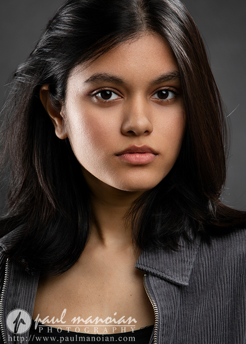 A young woman with straight, dark hair and brown eyes looks directly at the camera with a neutral expression. Photographed by an Ann Arbor Actor Headshots Photographer, she wears a gray zip-up jacket against a dark, plain background.