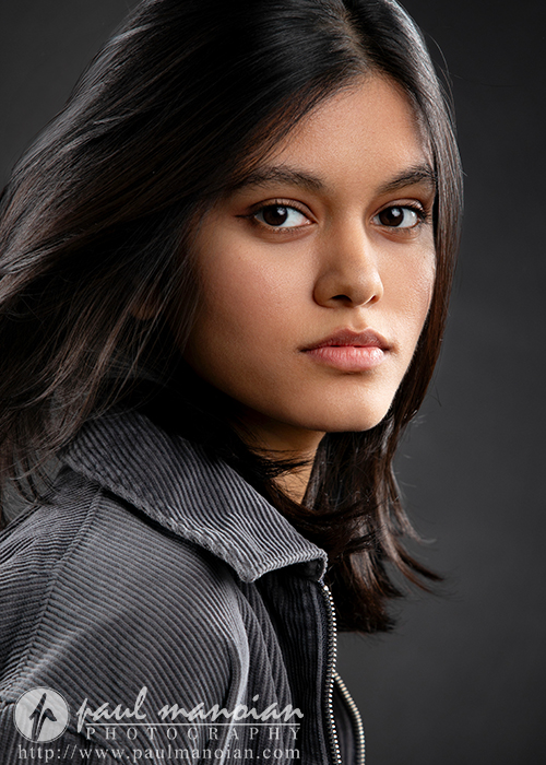 A young woman with straight dark hair and brown eyes looks into the camera. She wears a grey corduroy jacket and stands against a dark background. The image features the watermark “Paul Mangian Photography, Ann Arbor Actor Headshots Photographer.”.