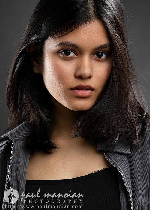 A young woman with medium-length dark hair and brown eyes looks directly at the camera. She wears a black top and a gray pinstripe jacket. Captured by an Ann Arbor Actor Headshots Photographer, the photo has a professional studio feel.