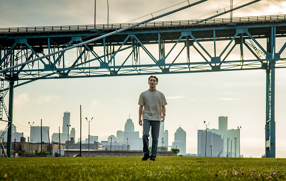 A high school in a light shirt and jeans walks on a grassy field beneath a large steel bridge, with a city skyline and cloudy sky in the background—perfect for Detroit senior pictures.