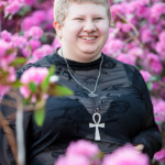 Person with short hair and black top smiles in front of bright pink flowers, wearing an ankh necklace—capturing a glimpse of their vibrant personality in these Ann Arbor senior pictures.