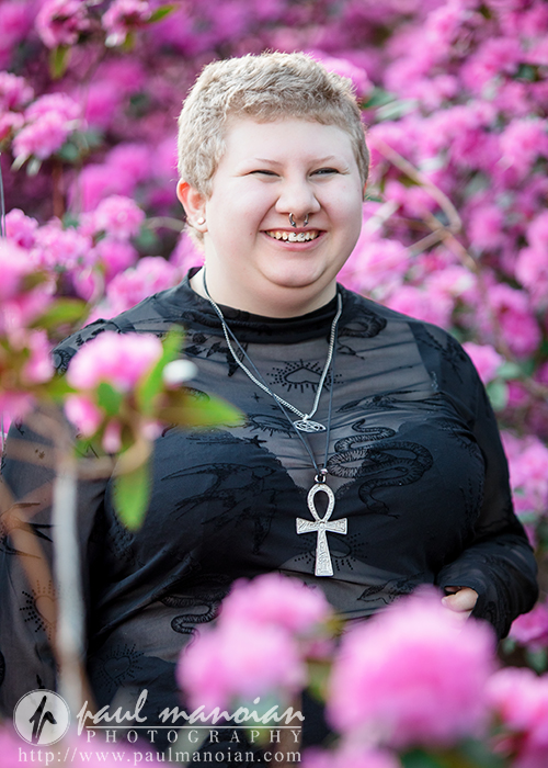 Person with short hair and black top smiles in front of bright pink flowers, wearing an ankh necklace—capturing a glimpse of their vibrant personality in these Ann Arbor senior pictures.