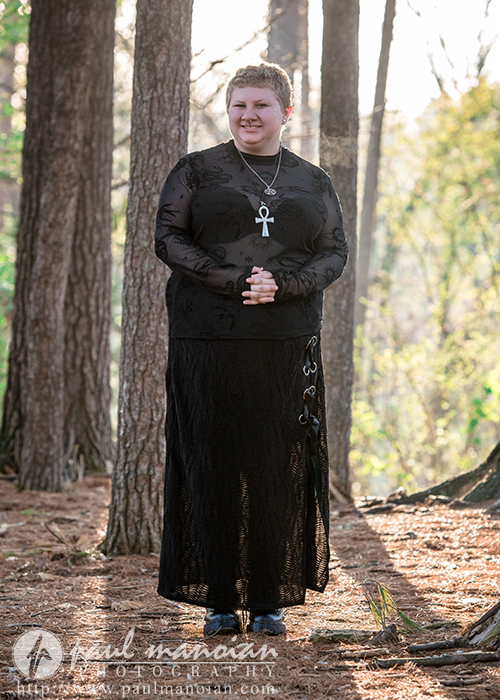 Person with short hair in a black outfit and ankh necklace stands in a sunlit Ann Arbor forest, smiling and letting their personality shine—perfect for senior pictures.