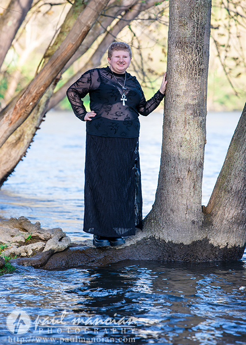 Woman in a long black lace dress stands by a tree near a river in Ann Arbor, her personality shining as she smiles at the camera—perfect for senior pictures.