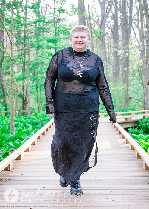 Confident personality shines as a person with short hair in a black sheer outfit walks on a wooden path in a lush green forest, perfect for unique senior pictures.