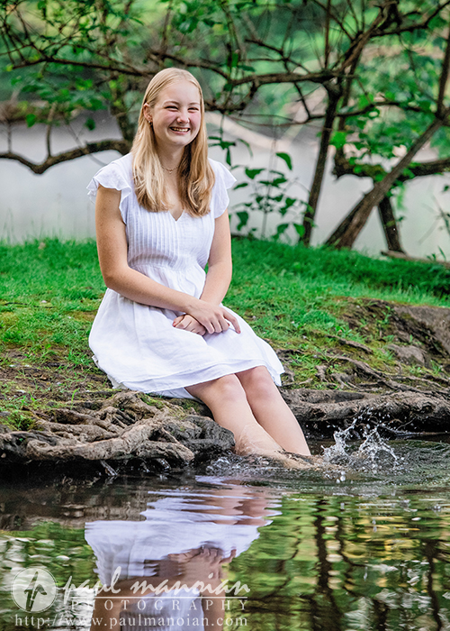 Smiling young woman in a white dress sits by a pond, splashing water with her feet, surrounded by greenery—a perfect moment for Ann Arbor senior pictures.
