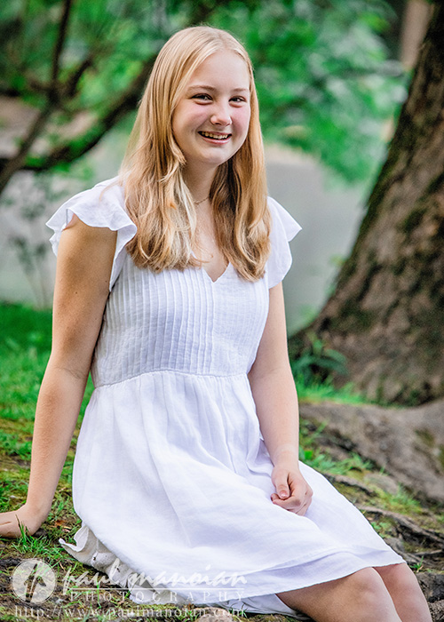 Smiling young woman in a white dress sits on grass beside a tree in a green outdoor setting, capturing the charm of Ann Arbor senior pictures.