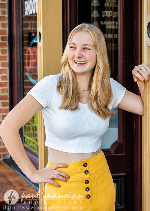 Teen girl with blonde hair in a white top and yellow skirt, smiling and leaning against a storefront doorway—perfect for stylish Ann Arbor senior pictures.