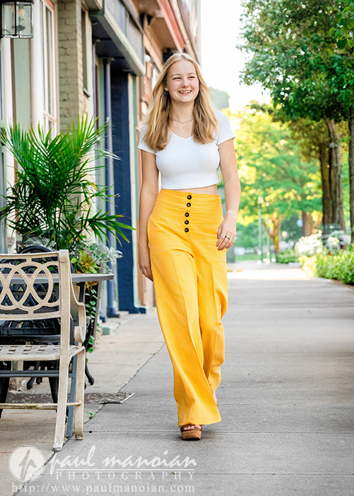 Smiling woman in yellow pants and white top walking on a sunny Ann Arbor sidewalk lined with plants and shops, perfect for vibrant senior pictures.
