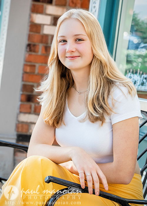 Teen girl with long blonde hair, white top, and yellow pants sitting outdoors, smiling at the camera during her Ann Arbor senior pictures.