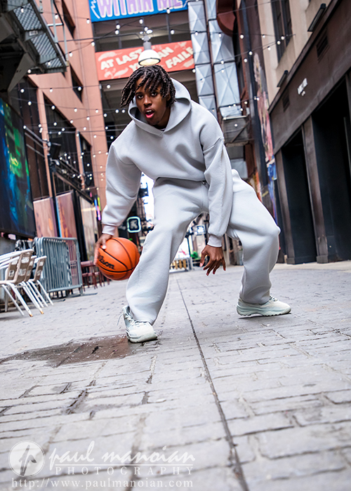 High school senior in a gray hoodie dribbling a basketball on a city alley with string lights overhead, perfect for capturing the urban vibe of Detroit senior pictures or creative downtown Detroit photography.