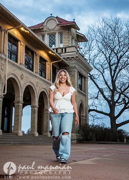 A smiling high school senior in ripped jeans and a white top walks outside a large historic building on a cloudy day, capturing her Belle Isle senior pictures session.