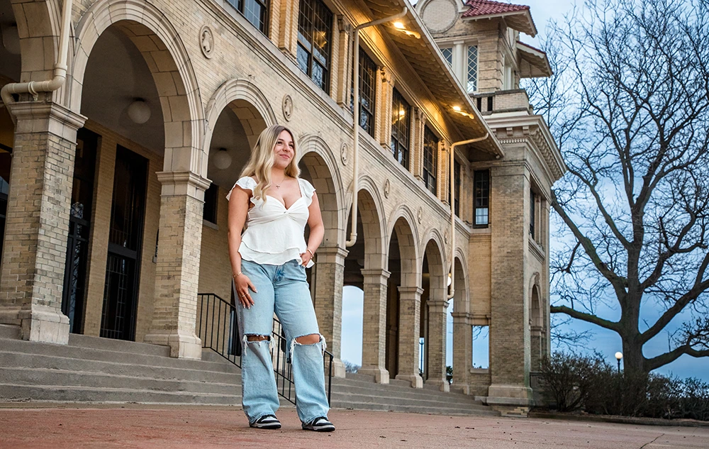 High school senior in ripped jeans and white top stands confidently in front of a historic building with arches for her Belle Isle senior pictures session.