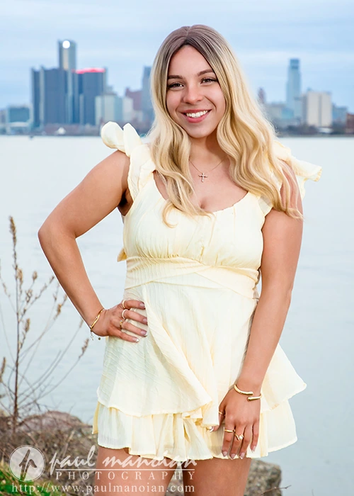 High school senior in a pale yellow dress posing by a waterfront with a city skyline behind her, smiling at the camera.