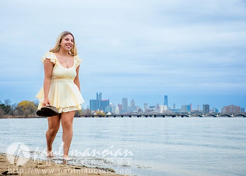 Smiling high school senior in a yellow dress walks barefoot by the water during her Belle Isle senior pictures session, with a city skyline in the background.