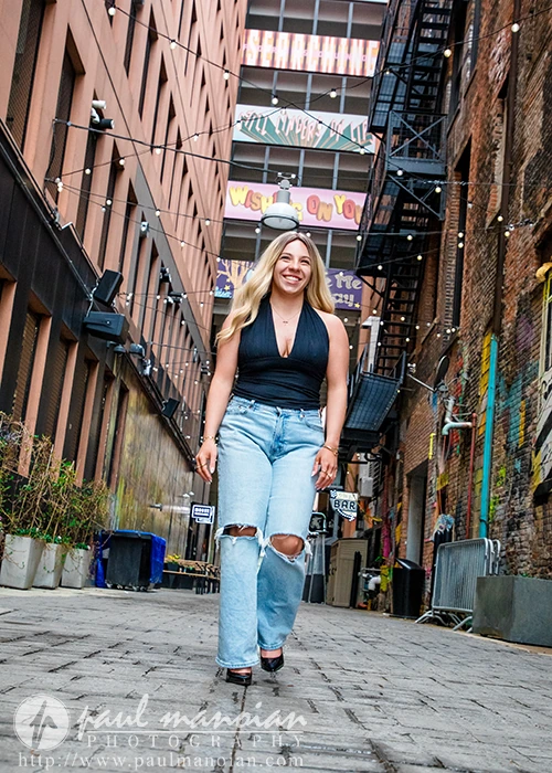High school senior in ripped jeans and black top smiles while walking through a vibrant Detroit alley, surrounded by murals and string lights for her senior pictures session
