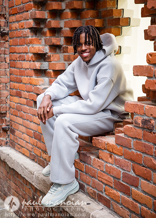 A high school senior in a gray tracksuit sits and smiles on a brick window ledge, capturing cool Detroit vibes for their senior pictures.