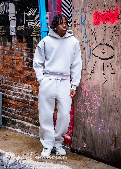 High school senior in a white hoodie and sweatpants stands in a graffiti-covered alley next to a wooden door, perfect for downtown Detroit senior photos with an edgy urban vibe.