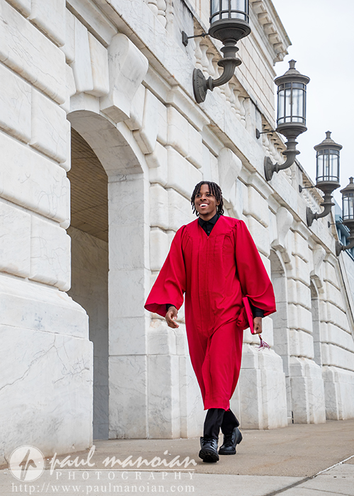 High school graduate in a red cap-and-gown walking along a stone colonnade with ornate lanterns outside a historic building
