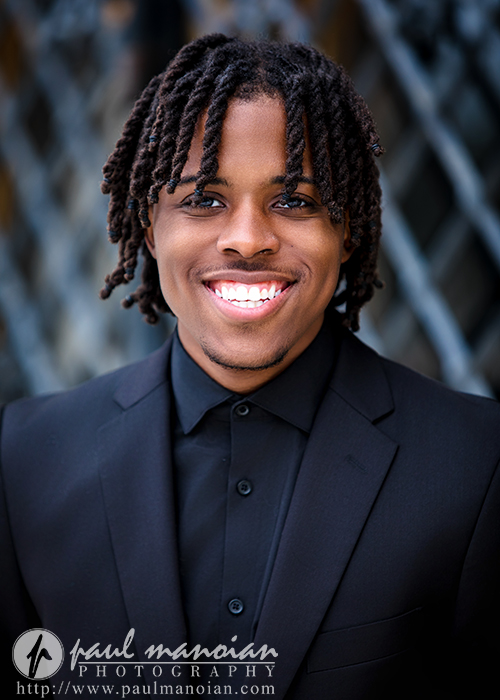 Portrait of a smiling high school senior with dreadlocks wearing a black suit, outdoors with a blurred background.