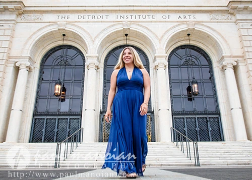 A high school senior in a blue dress stands smiling on the steps outside the Detroit Institute of Arts, capturing her senior pictures at this iconic location.