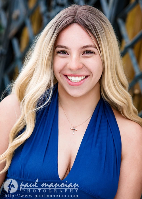 Smiling high school senior with long blonde hair, wearing a blue dress and a cross necklace, outdoors for senior pictures at the Detroit Institute of Arts.