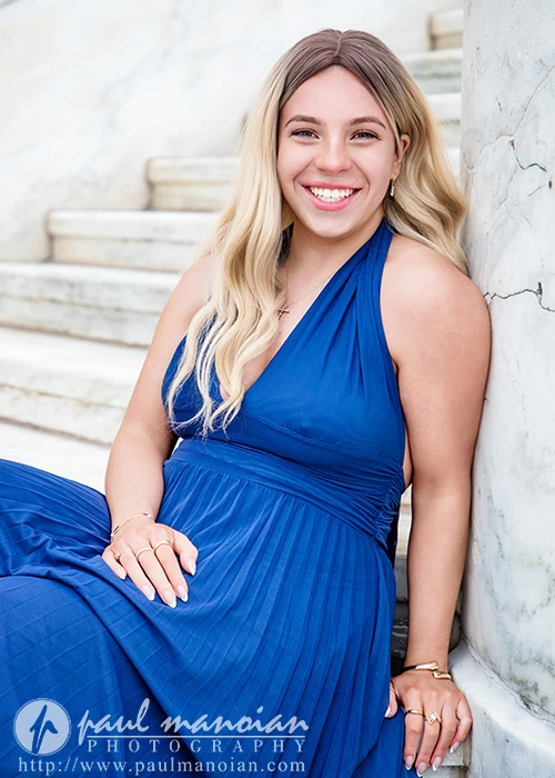High school senior with long blonde hair in a blue dress sitting on marble steps, smiling at the camera during her senior pictures at the Detroit Institute of Arts.