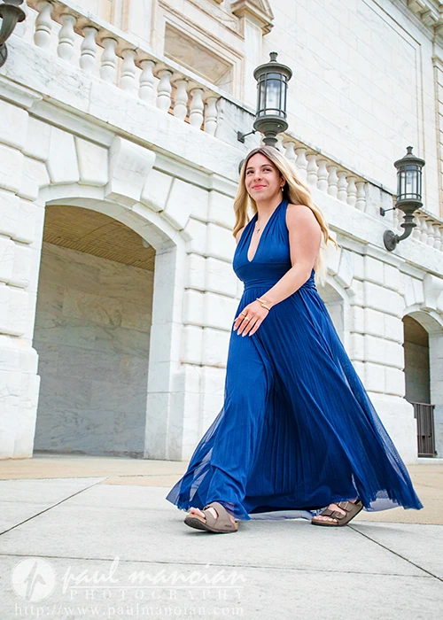 High school senior in a flowing royal blue gown walking on a stone plaza beside a marble building with arches and lanterns in the background, facing the camera.