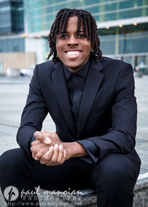 Smiling high school senior in a dark suit sits on concrete steps outside a glass-fronted building, looking at the camera.