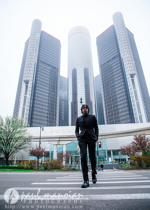 A high school senior in a black suit walks on a city street in front of tall, modern glass buildings, capturing the urban vibe perfect for senior pictures Detroit style.