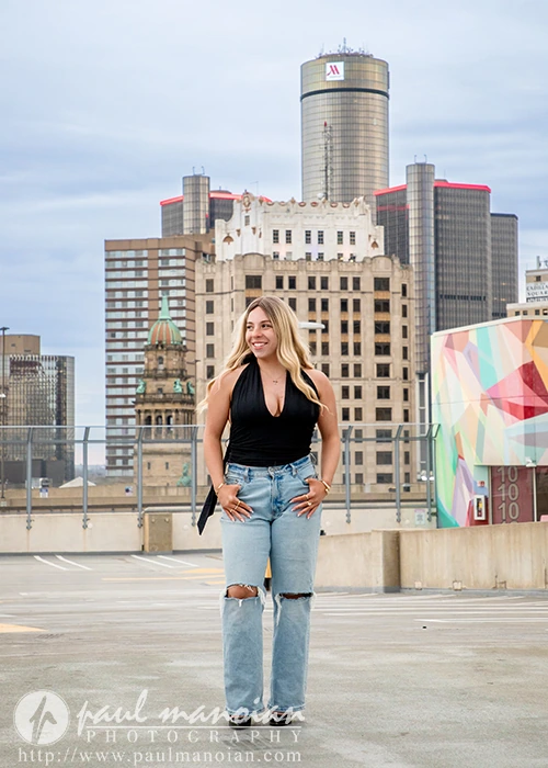 High school senior in ripped jeans and black top standing on a Detroit rooftop, city skyscrapers in the background for her senior pictures session