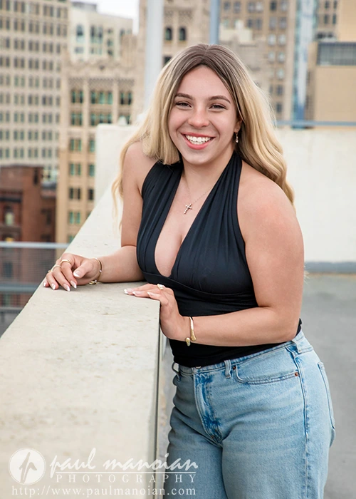 High school senior with blonde hair in a black halter top and jeans smiling on a Detroit rooftop, city buildings behind her for a senior pictures session