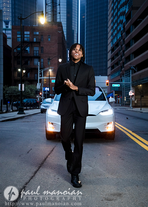A high school senior in a black suit stands smiling in front of a white car on a city street at dusk, capturing the essence of downtown Detroit senior photos.