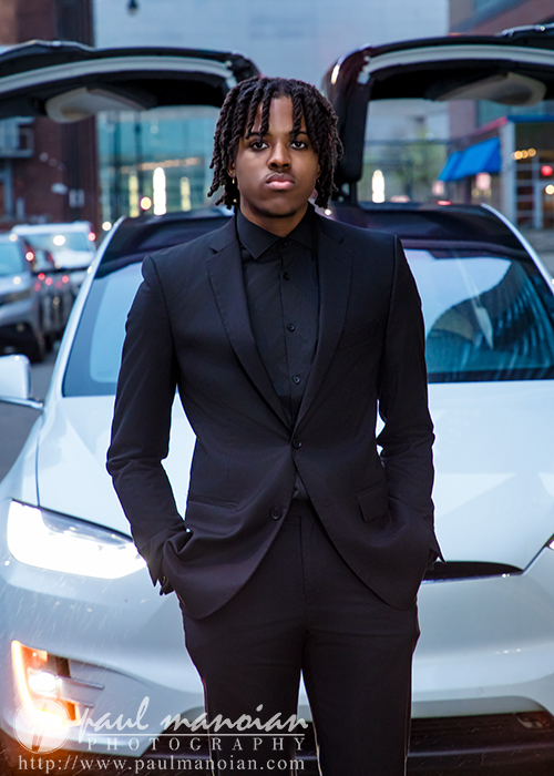 A high school senior in a black suit stands confidently in front of a white Tesla with its doors open during a downtown editorial session on a bustling city street.