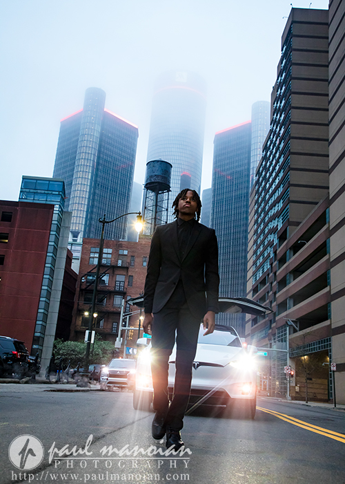 A high school senior in a dark suit walks on a city street with tall buildings and a Tesla with headlights behind him, perfect for downtown Detroit senior photos or unique senior pictures with car.
