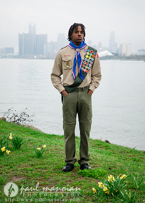A high school senior Eagle Scout stands on grass by a lake with a city skyline in the background, wearing a uniform and merit badge sash—perfect for downtown Detroit photography sessions.
