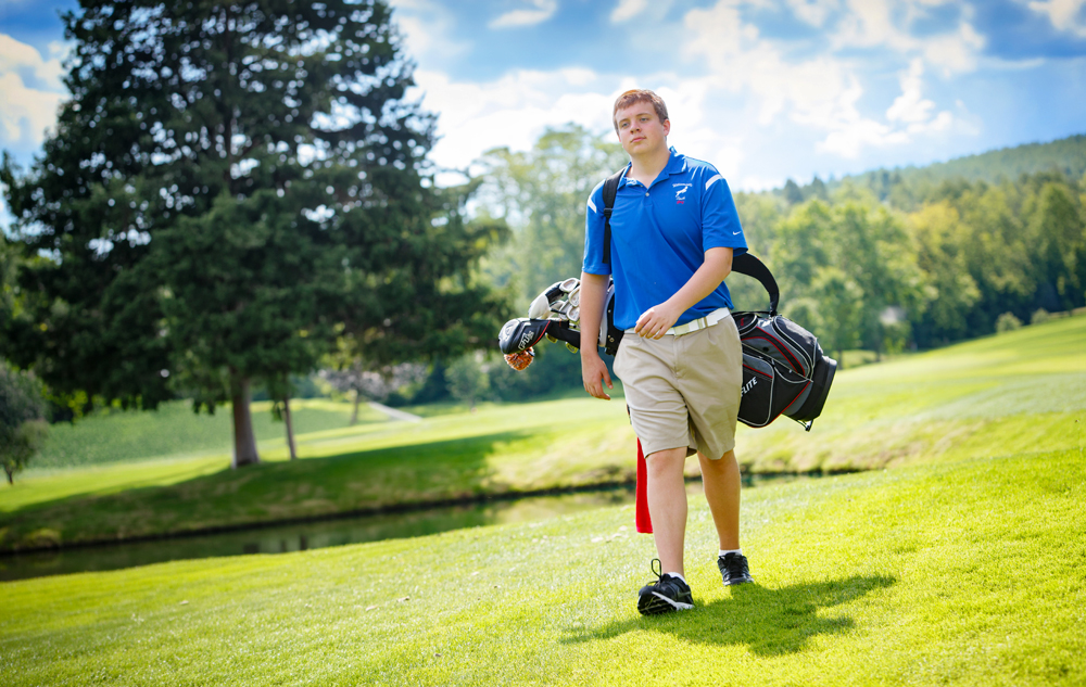 A high school senior walks on a sunny golf course carrying a golf bag over his shoulder, capturing the perfect moment for golf senior pictures.