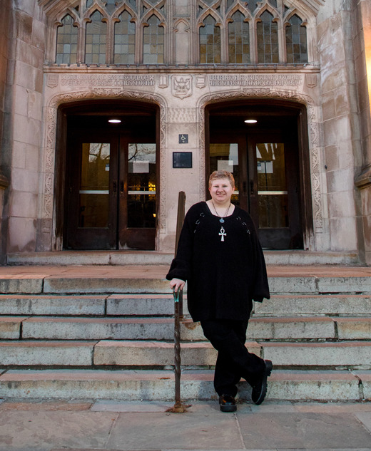 A high school senior in black clothing stands on stone steps outside a building with large wooden doors and glowing lanterns, capturing a moment full of personality—perfect for senior pictures in charming Ann Arbor.