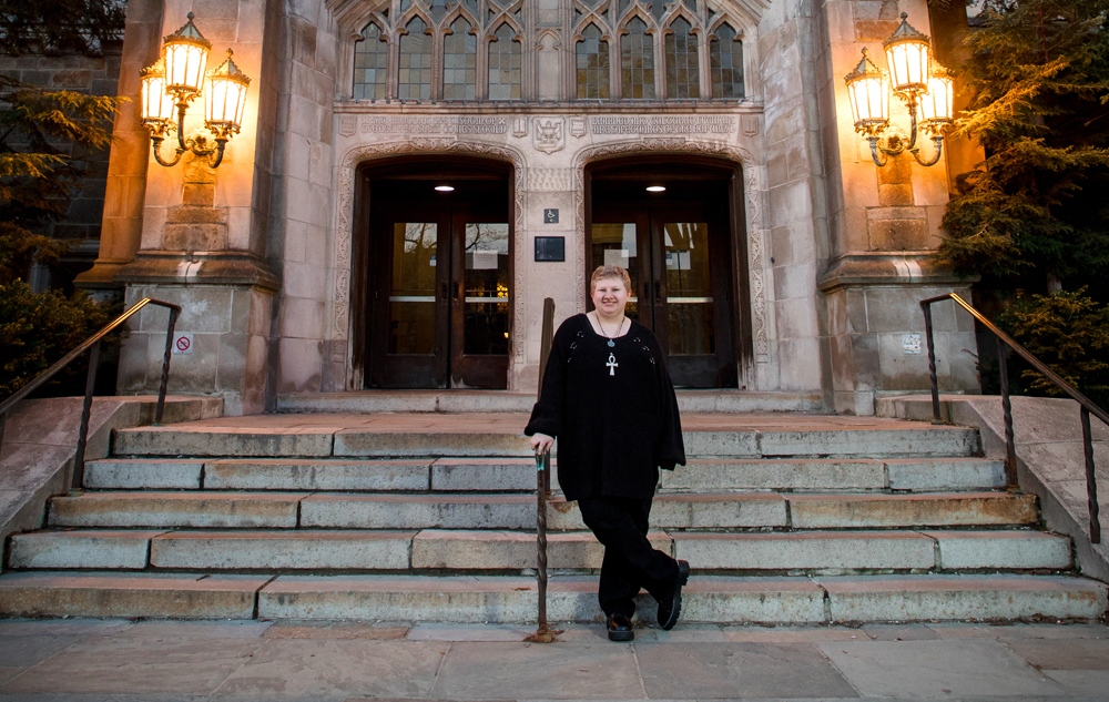 A high school senior in black clothing stands on stone steps outside a building with large wooden doors and glowing lanterns, capturing a moment full of personality—perfect for senior pictures in charming Ann Arbor.