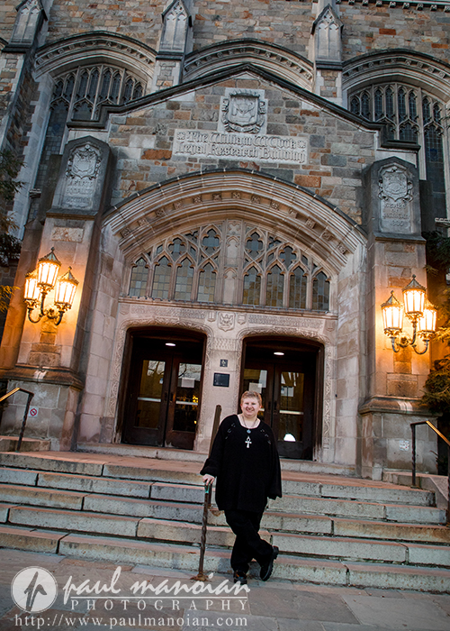 A person in black, radiating personality, stands smiling on stone steps outside a large, ornate Ann Arbor building with arched doorways and lamps—perfect for senior pictures.