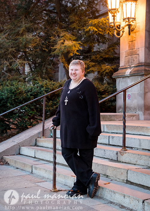 Person in black clothing with a cane stands on outdoor steps near a lamp post in Ann Arbor, smiling at the camera, showing lots of personality.