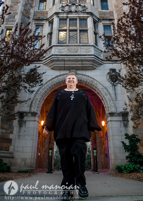 Person in a black sweater walks confidently under a stone archway of a historic Ann Arbor building with lit lamps, capturing personality perfect for senior pictures.