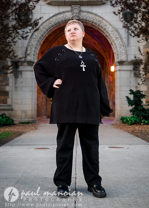 Person with short hair in black clothing stands confidently on a sidewalk in front of a stone archway, showcasing their bold personality—perfect inspiration for senior pictures in Ann Arbor.