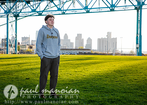 High school senior boy in a gray hoodie stands on grass under a bridge with the city skyline behind him, captured during a sunrise session Detroit, perfect for unique senior pictures.