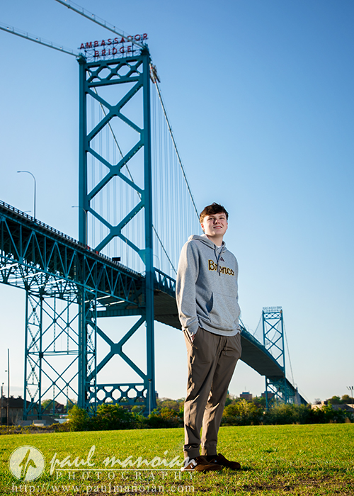 High school senior boy in casual clothes stands on grass near the blue Ambassador Bridge under a clear sky, capturing unique Senior Pictures Detroit.